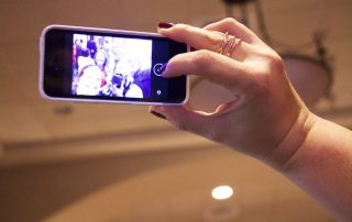 Hand die ein Smartphone hält und gerade ein Selfie schießt mit nicht erkennbaren Gesichtern.